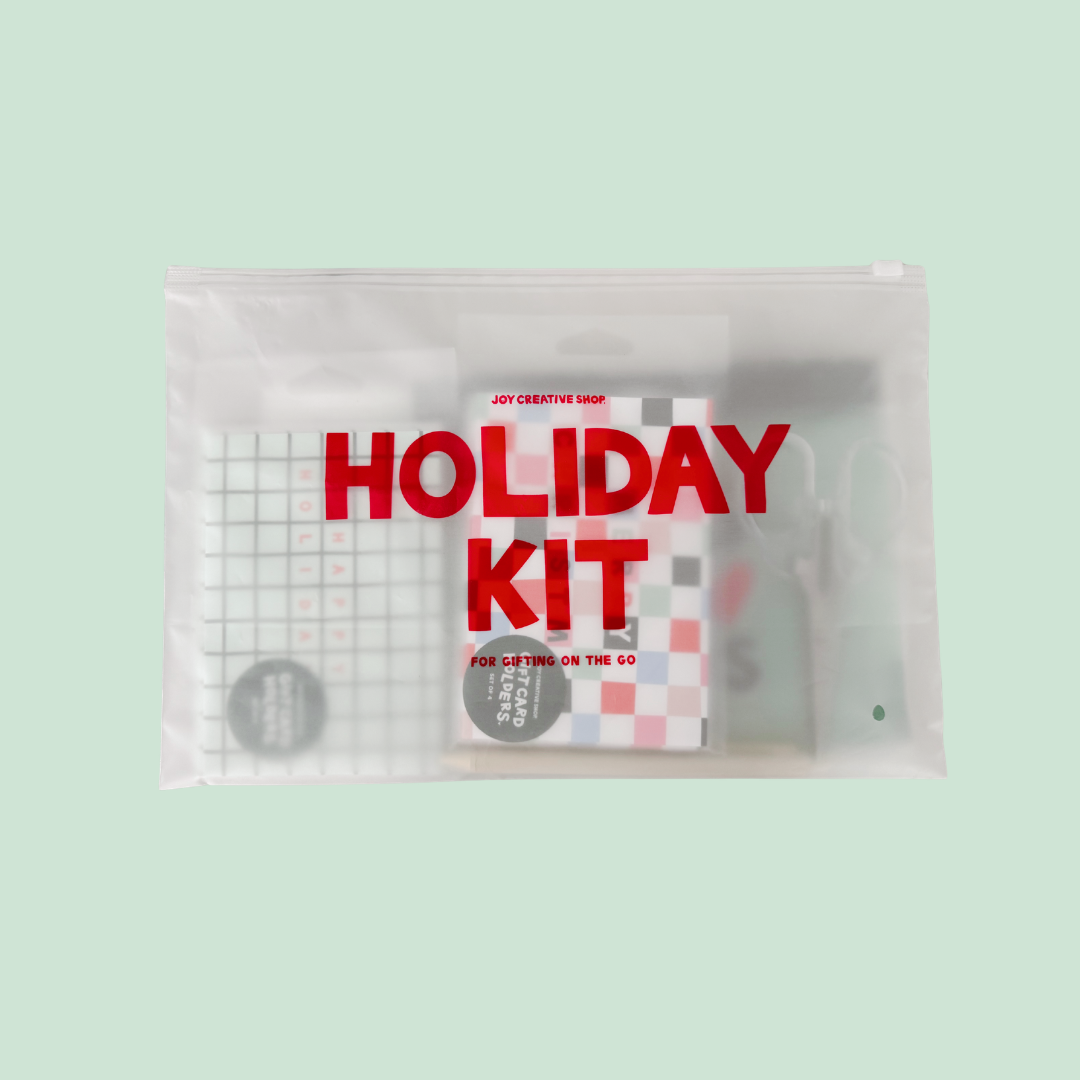 A clear zippered pouch labeled 'HOLIDAY KIT' in large red text, containing assorted small items including a checkered notebook, a black circular item, and other accessories, set against a pale mint green background.